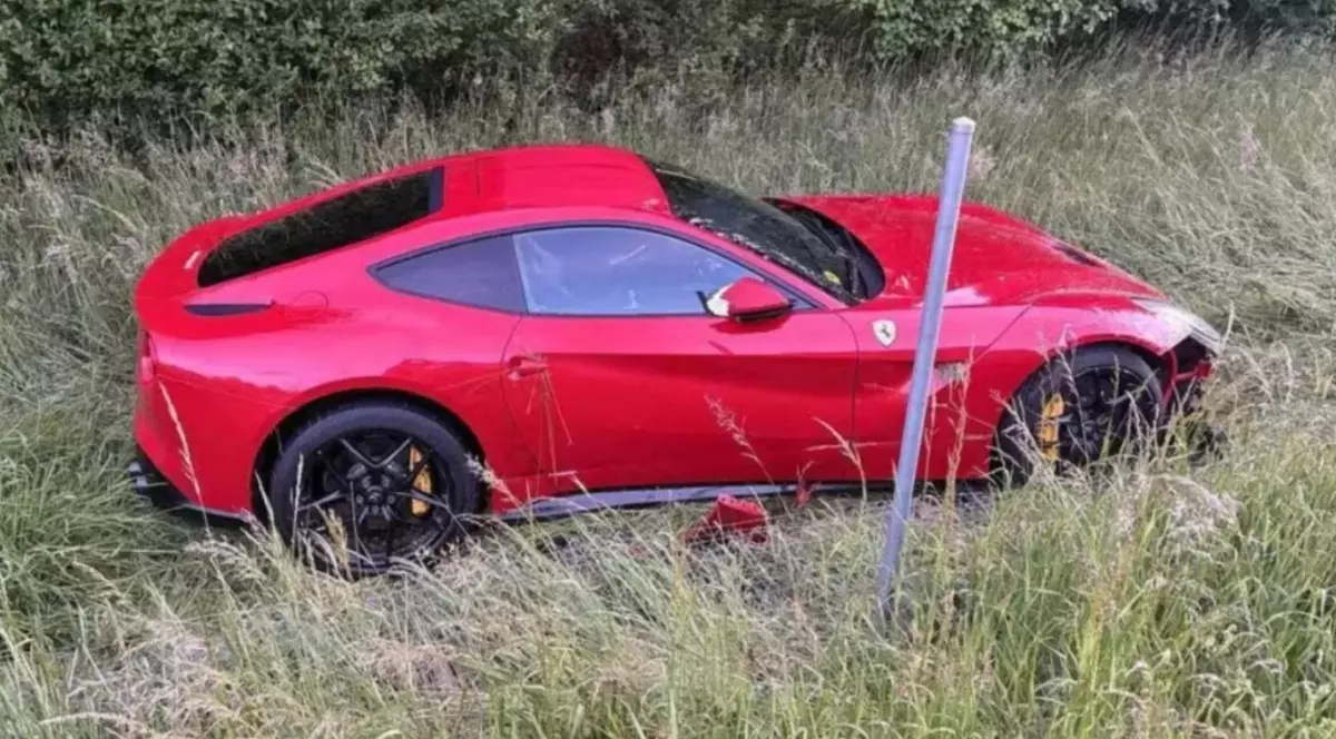 Imaginea prezintă un autoturism sport marca Ferrari, de culoare roșie, care a părăsit carosabilul și s-a oprit într-un șanț cu iarbă înaltă de pe marginea drumului.