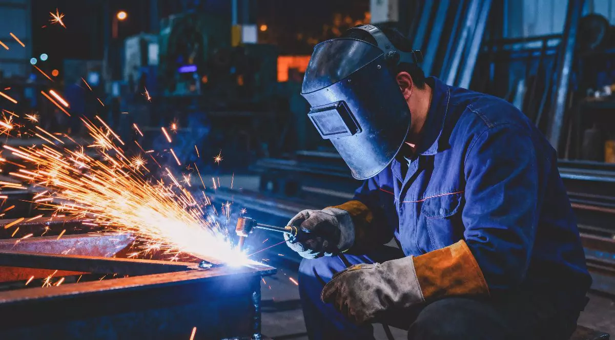Imaginea surprinde un sudor echipat cu mască de protecție și mănuși speciale în timp ce lucrează la o structură metalică, generând scântei luminoase în atelierul industrial.