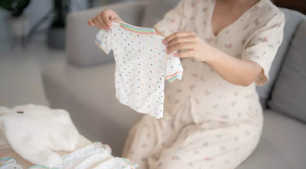 Imaginea surprinde o mamă care pregătește haine pentru bebeluș, ținând în mâini un body alb pentru copii. Fotografia ilustrează grija pentru alegerea hainelor celor mici și se potrivește unui articol despre închirierea articolelor vestimentare pentru bebeluși și copii.