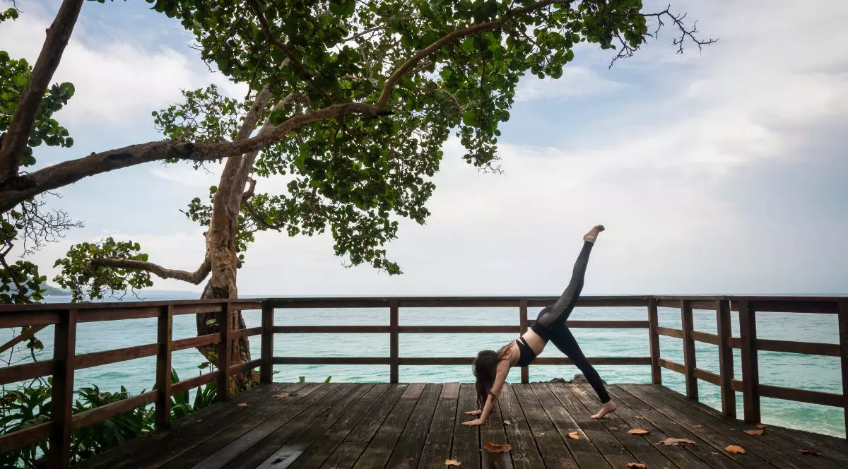 O scenă liniștită de wellness tropical, în care o femeie practică yoga pe o terasă din lemn situată pe malul oceanului, înconjurată de copaci verzi luxurianți și cu vedere spre mare.