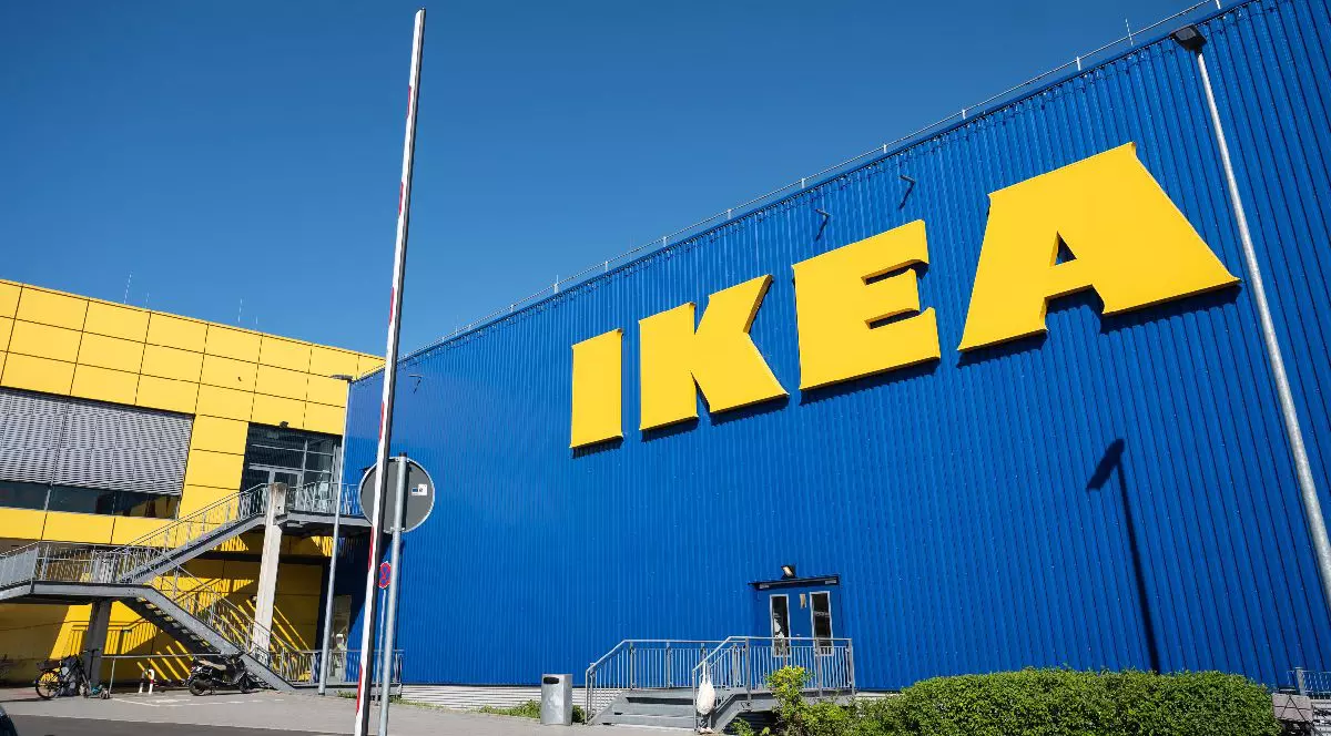 Imaginea surprinde o vedere laterală a unui magazin IKEA, cu fațata albastră și litere tridimensionale mari, galbene.