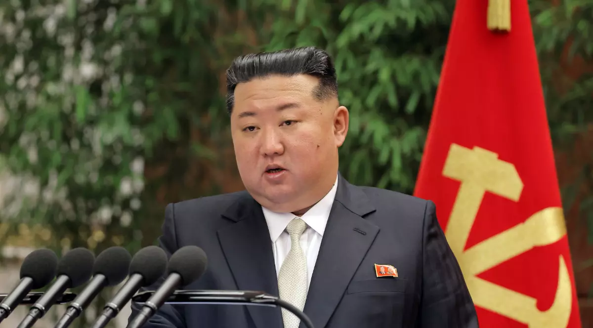 Kim Jong-un apare la un pupitru oficial, în timpul unui discurs susținut în fața mai multor microfoane. În spatele liderului nord-coreean se vede steagul Partidului Muncitorilor din Coreea, iar cadrul sugerează o reuniune sau un eveniment politic oficial.