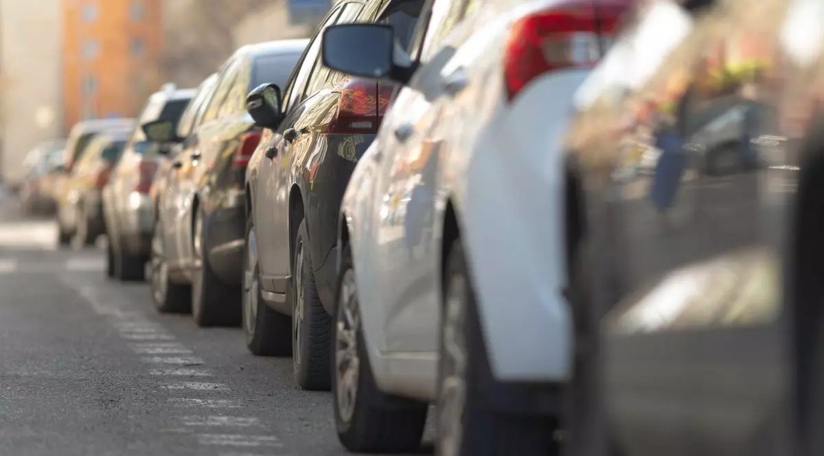 Mai multe mașini, albe, negre și gri, sunt într-o parcare pe stradă și sunt puse una în spatele alteia