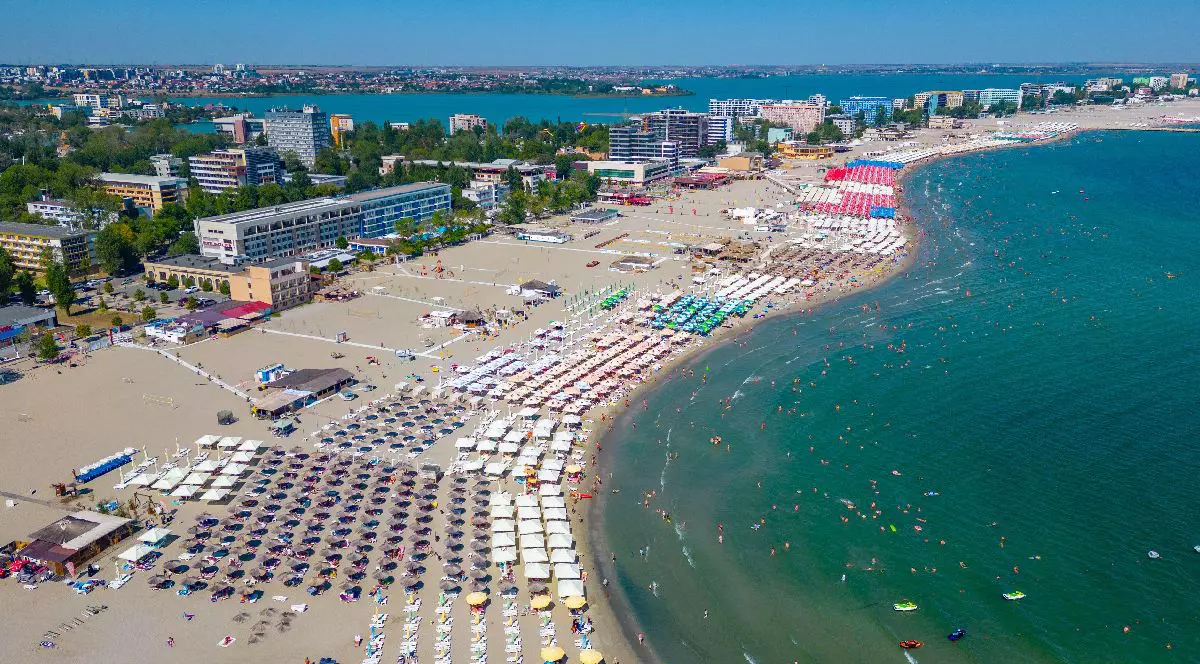 Imaginea prezintă o vedere aeriană a unei plaje aglomerate de pe litoralul românesc, cu numeroase umbrele colorate, hoteluri în fundal și turiști sub un cer senin.