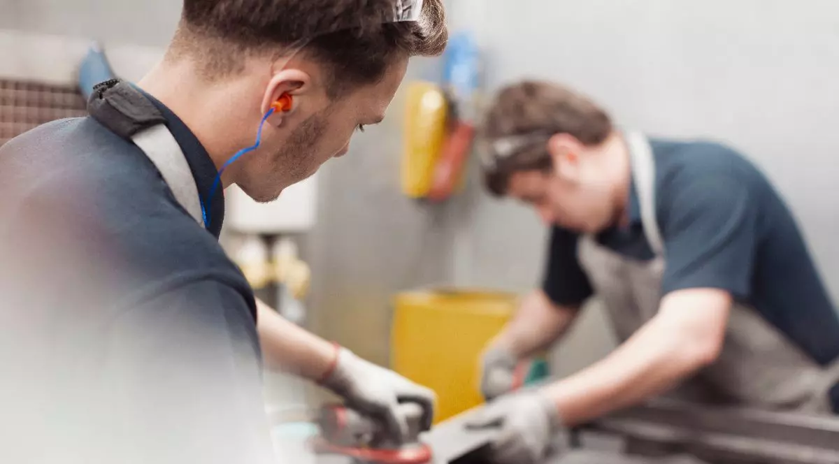 Imaginea surprinde doi muncitori u echipament de protecție în timp ce prelucrează cu atenție piese metalice într-un atelier industrial.
