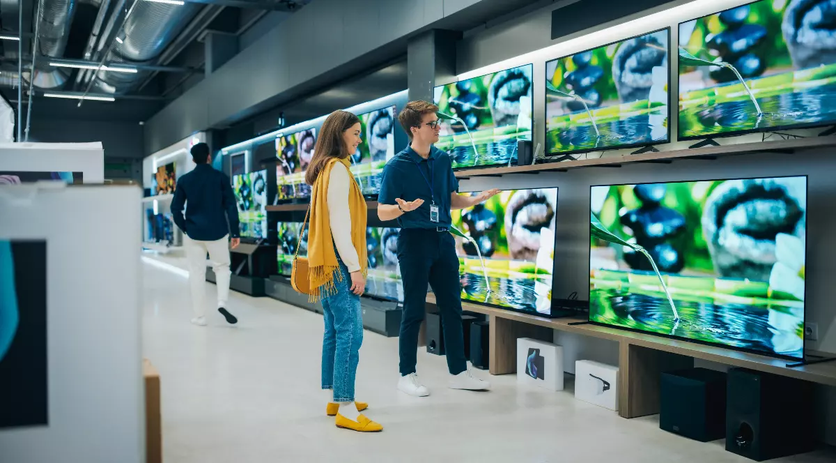 Imaginea surprinde interiorul unui magazin de electronice, unde o clientă analizează mai multe televizoare expuse pe rafturi, în timp ce un angajat îi oferă explicații despre produse. Fotografia ilustrează perioada campaniilor de reduceri și interesul crescut pentru electronice și electrocasn