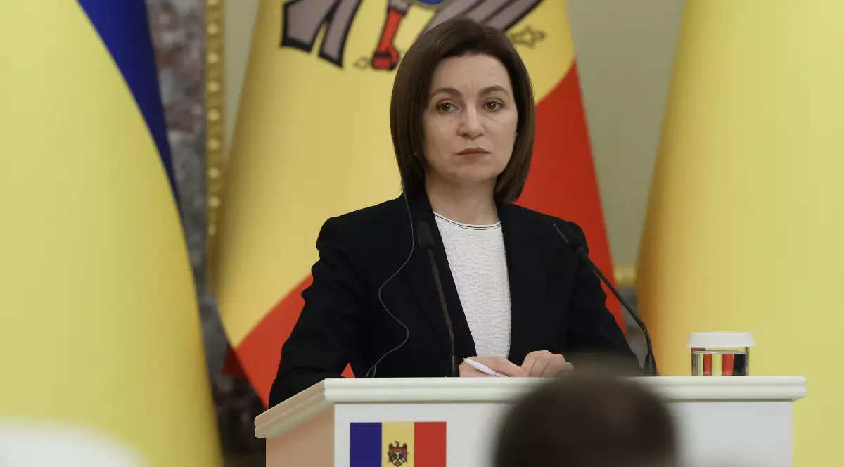 Imaginea o prezintă pe Maia Sandu vorbind de la o tribună oficială decorată cu steagul Republicii Moldova, având în fundal drapelul de stat parțial vizibil.