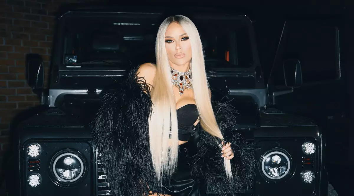 Artista apare într-o ținută all black elegantă, cu părul lung și blond, pozând în fața unei mașini de lux negre. Imaginea transmite opulență și un stil extravagant, completat de accesoriile strălucitoare și atitudinea încrezătoare.