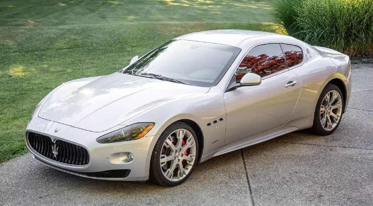 O imagine elegantă prezintă un Maserati GranTurismo argintiu parcat pe o alee cimentată, evidențiind designul și emblema distinctivă a tridentului pe grila frontală, într-un decor cu gazon verde.