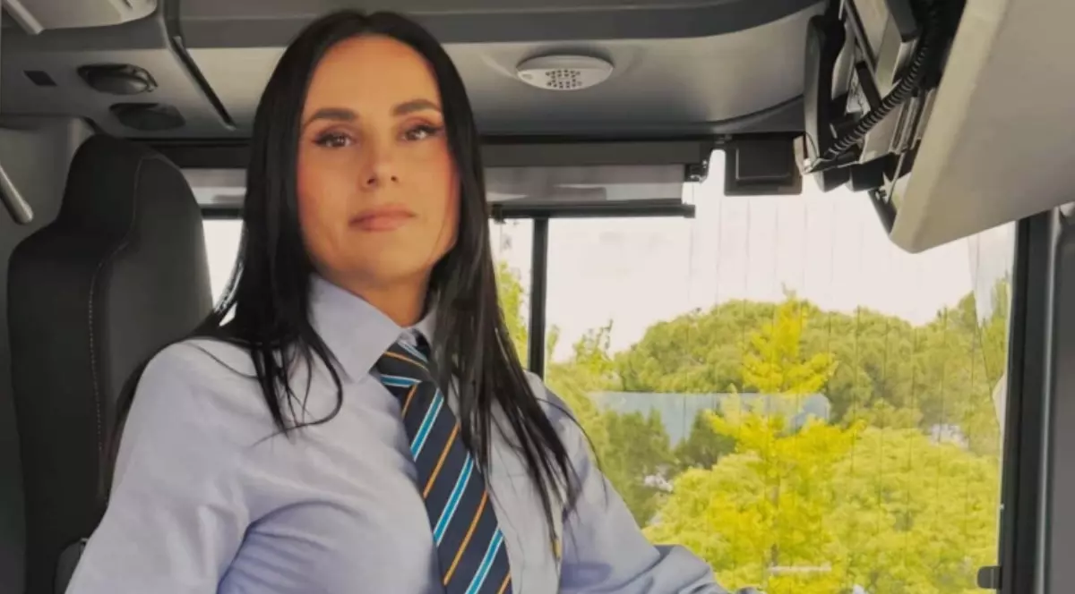 O femeie brunetă în cămașă, la volanul unui autobuz.