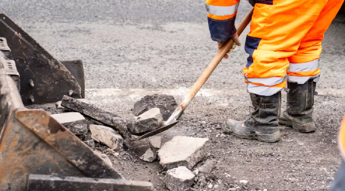 Un bărbat într-o salopetă portocalie folosește o lopată pentru a curăța un drum.
