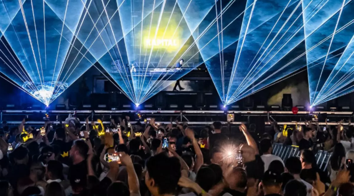 Imaginea surprinde atmosfera vibrantă a unui festival de muzică, cu sute de oameni adunați în fața scenei, în timp ce jocurile de lumini și lasere creează un spectacol impresionant. În centrul scenei apare logo-ul KAPITAL, iar publicul filmează și dansează pe ritmurile muzicii electronice.