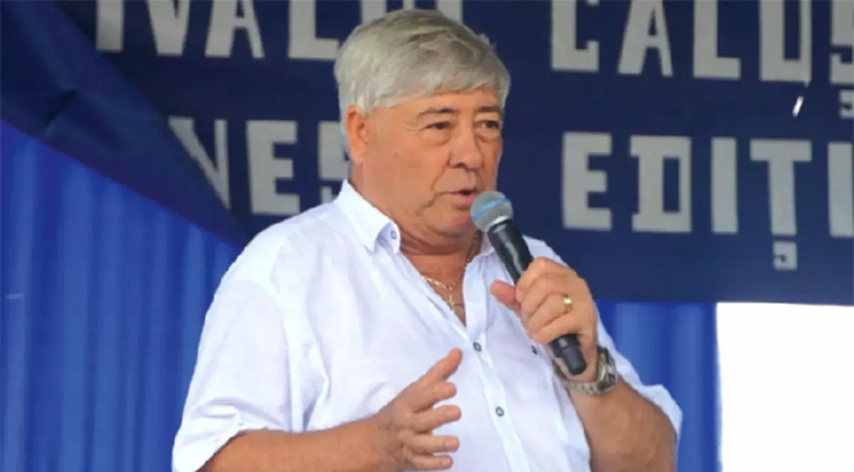 Imaginea îl surprinde pe Nicolae Stan în timpul unui discurs public, ținând un microfon în mână și adresându-se audienței de pe o scenă. Acesta poartă o cămașă albă și apare pe fundalul unui panou al unui eveniment organizat în aer liber.