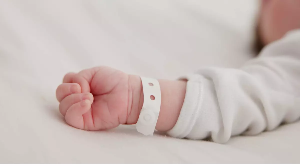 Mâna unui bebeluș, cu o brățară de spital, întinsă pe un pat cu lenjerie albă.