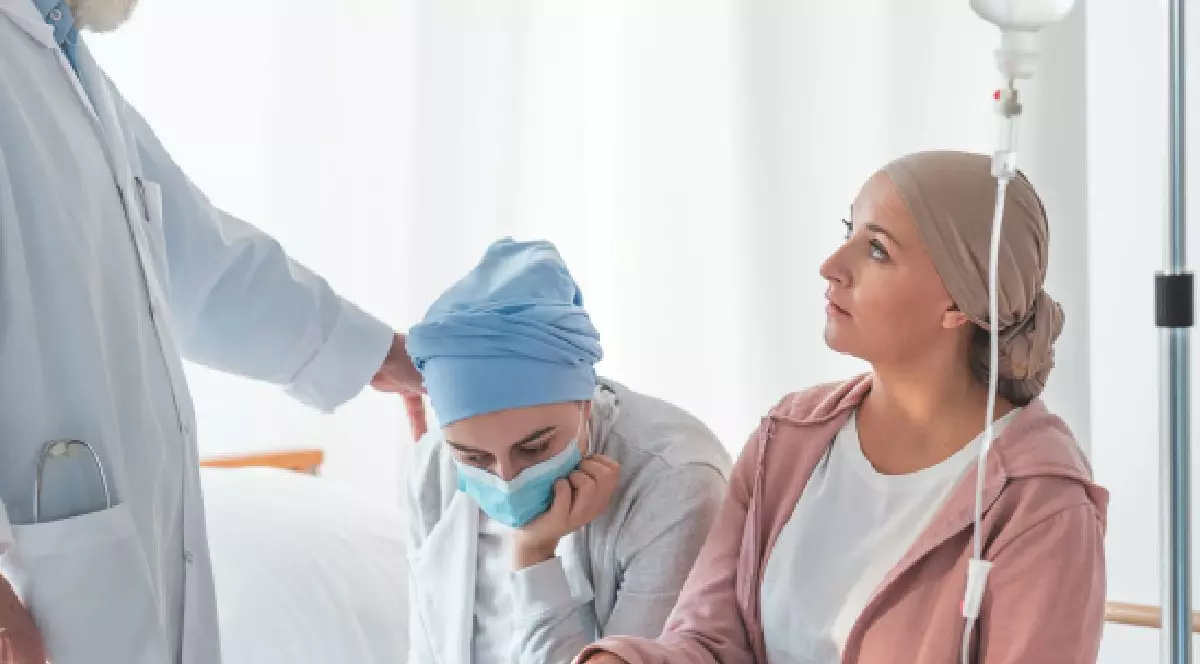 Intr-un salon sunt doua femei cu batic pe cap , una dintre ele cu masca, si cu o perfuzie pusa pe stativ, iar o persoana in halat alb ii pune o mana pe umar uneia dintre femei. leucemie transplant medular, centre supraaglomerate