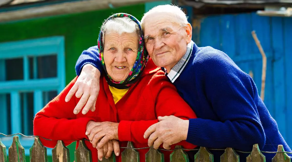 Un prim-plan cu doi pensionari cu zâmbetul pe buze, pe veranda unei case de la țară. Femeia, îmbrăcată cu un pulover roșu, este ținută de gât de soț, îmbrăcat cu cămașă și un pulover albastru