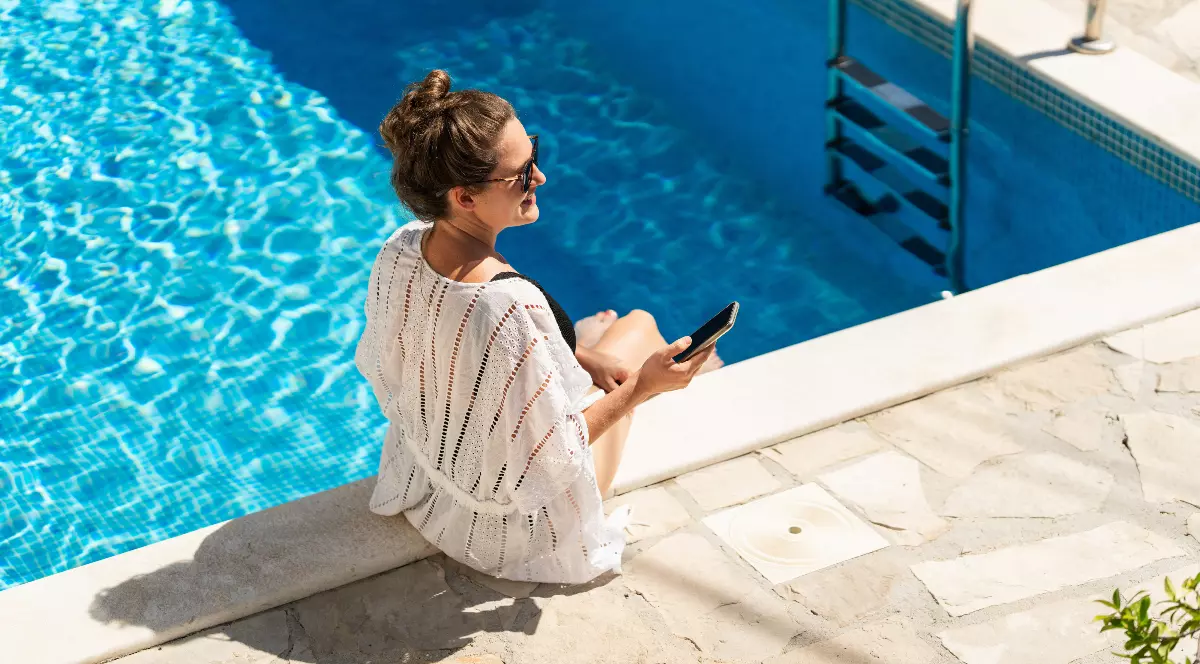 O femeie stă relaxată la marginea unei piscine cu apă limpede, ținând telefonul în mână și bucurându-se de vremea însorită. Atmosfera transmite liniște și vacanță, iar decorul elegant completează perfect imaginea unei zile de vară petrecute la piscină.