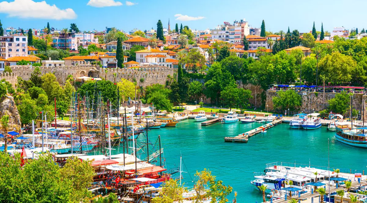 Această imagine surprinde pitorescul port din Antalya, o destinație turistică plină de viață de pe Riviera Turcească. Situat pe malul apelor turcoaz ale Mării Mediterane, acest oraș de coastă este renumit pentru îmbinarea dintre patrimoniul antic, stâncile spectaculoase și atmosfera modernă de litoral. Fotografia pune în evidență cartierul istoric Kaleiçi, care domină portul de agrement. Casele tradiționale cu acoperișuri din țiglă roșie se înalță deasupra fortificațiilor din piatră, în timp ce vegetația luxuriantă înconjoară zidurile vechi ale orașului. Mai jos, iahturile și bărcile de excursie se odihnesc în apele calme și limpezi ale portului, creând un cadru maritim pitoresc. Un dig curbat protejează portul de agrement, iar stâncile abrupte încadrează țărmul, oferind priveliști panoramice uimitoare.