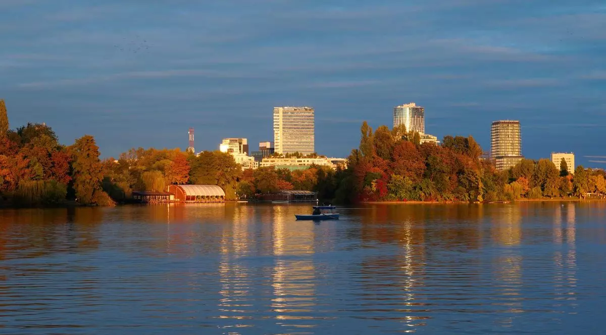 Peisaj frumos de toamnă la ora aurie în Parcul Herăstrău din București, cu bărci pe lac, copaci colorați și turnuri de birouri în depărtare
