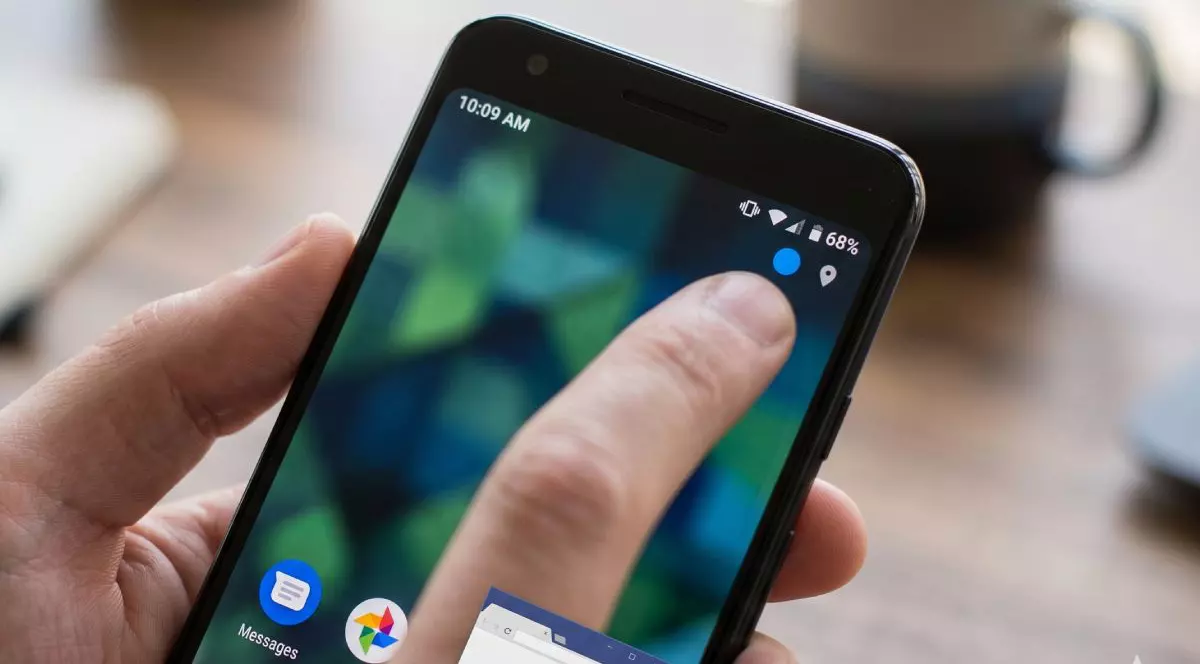 O persoană ține în mână un telefon mobil cu Android și indică cu un deget spre un punct albastru din colțul ecranului