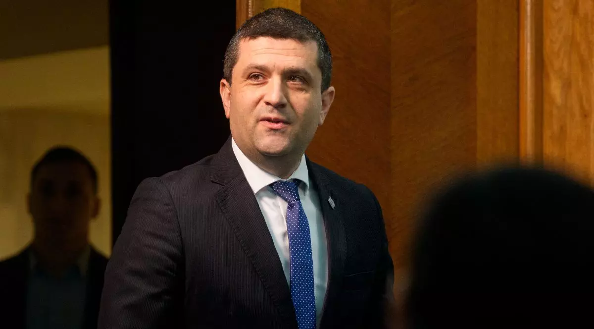 Radu Miruță, îmbrăcat în costum închis la culoare și cu cravată albastră ci buline mici albe, lângă o ușă din lemn masiv cu o persoană estompată în fundal