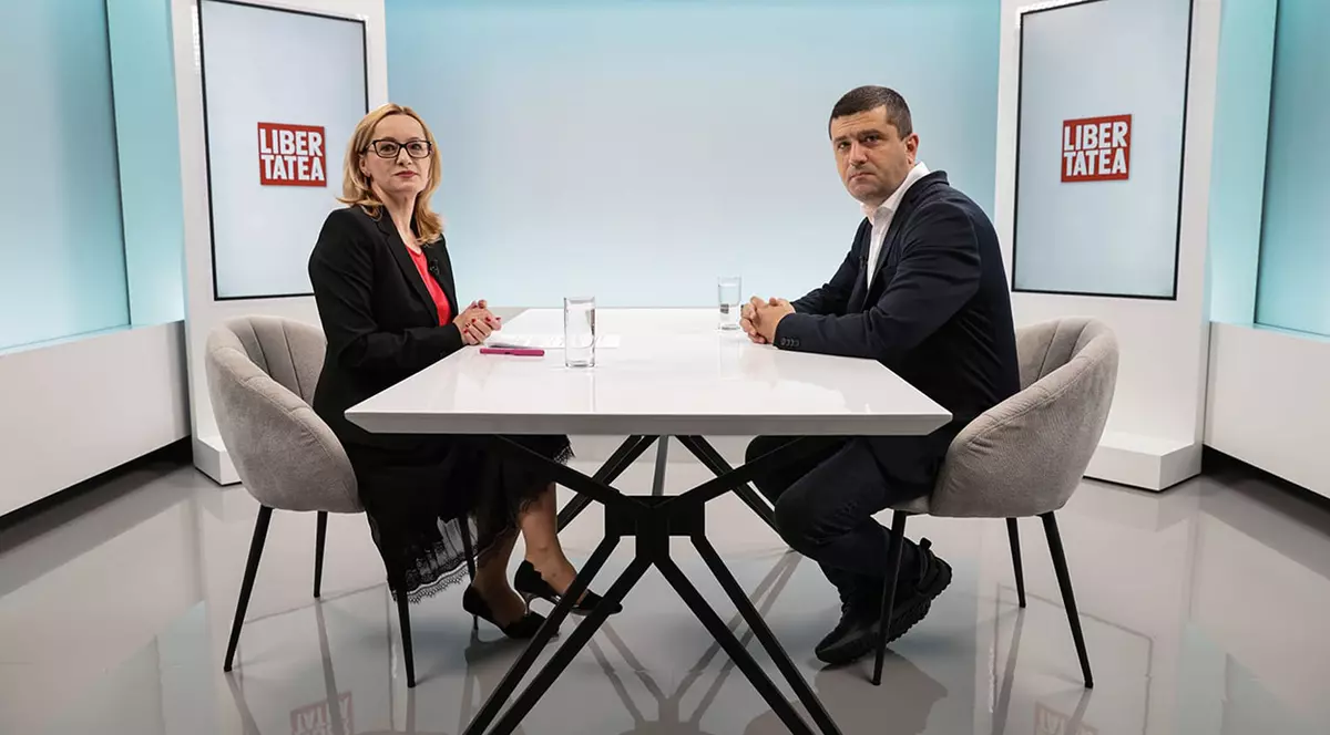 Anca Grădinaru, jurnalist Libertatea, îmbrăcată într-o ținută neagră cu bluză roșie față în față cu ministrul Radu Miruță îmbrăcat intr-un costum închis la culoare cu cămașă albă.