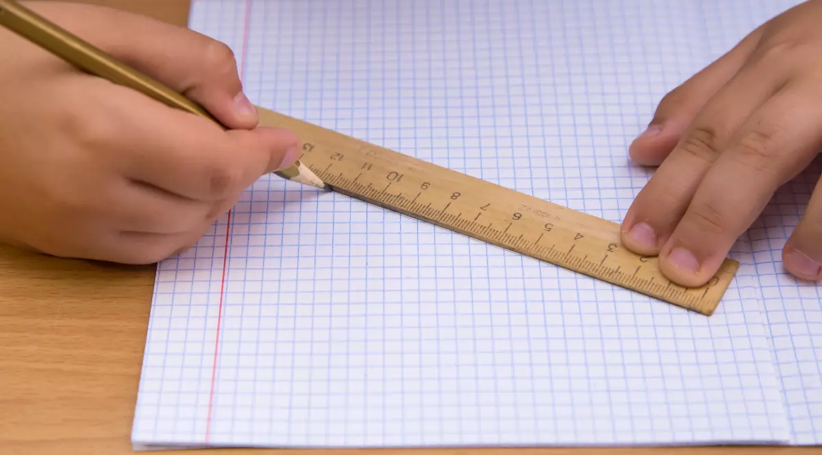 Un elev folosește o riglă din lemn pentru a trasa o linie pe o foaie de matematică cu pătrățele. Imaginea surprinde un moment obișnuit din timpul unei activități școlare, evidențiind utilizarea instrumentelor clasice de măsurare și desen.