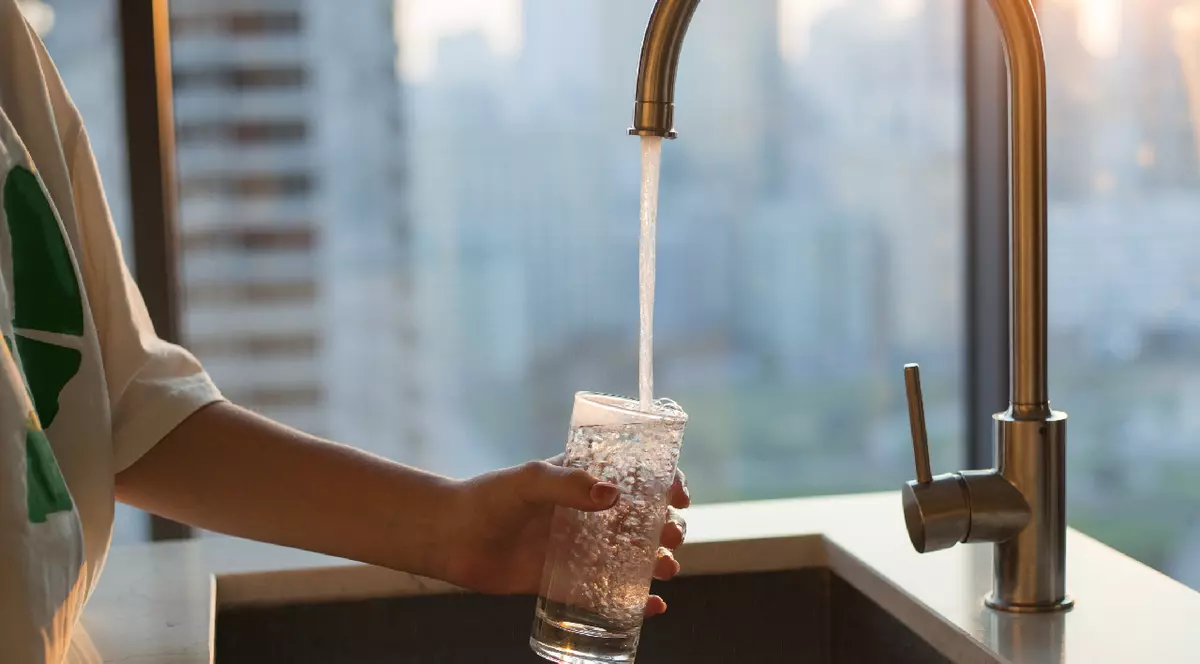 Fata umple un pahar cu apă de la robinet filtrată curată pentru băut în bucătărie acasă. Fereastră în fundal. Robinet modern și chiuvetă în bucătăria de acasă.
