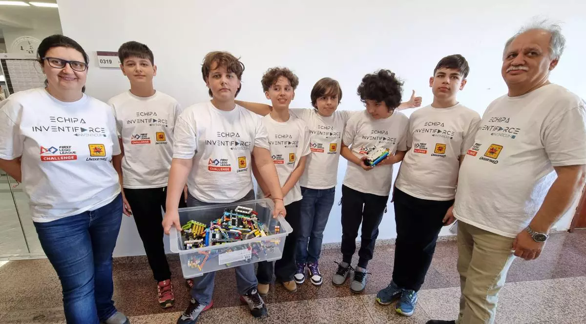 Șase elevi din Brașov , in tricouri albe cu inscriptii, reprezintă România la Asia Pacific Open Championship 2026, langa ei stanga dreapta mentorii coordonatori, un baiat tine ridicata o cutie cu lego