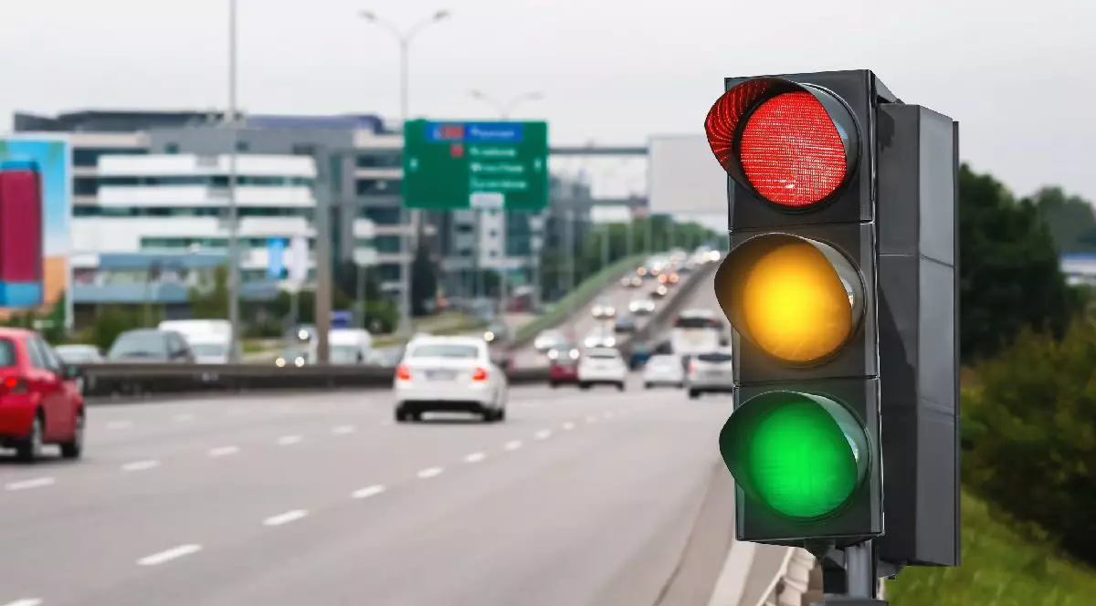 Imaginea prezintă în prim-plan un semafor rutier cu toate cele trei lumini -roșu, galben și verde - aprinse simultan, având în fundal o șosea aglomerată cu mașini aflate în trafic.