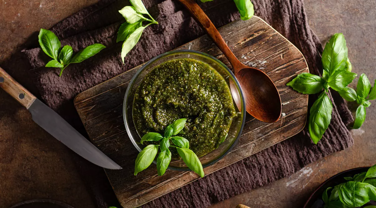 Secretul bucătarilor profesioniști: 3 sosuri de casă care schimbă complet gustul oricărui preparat - Imagine realizată de sus care prezintă un bol din sticlă transparentă umplut cu sos pesto de un verde intens, aşezat pe un fund de lemn. înconjurat de frunze proaspete de busuioc.