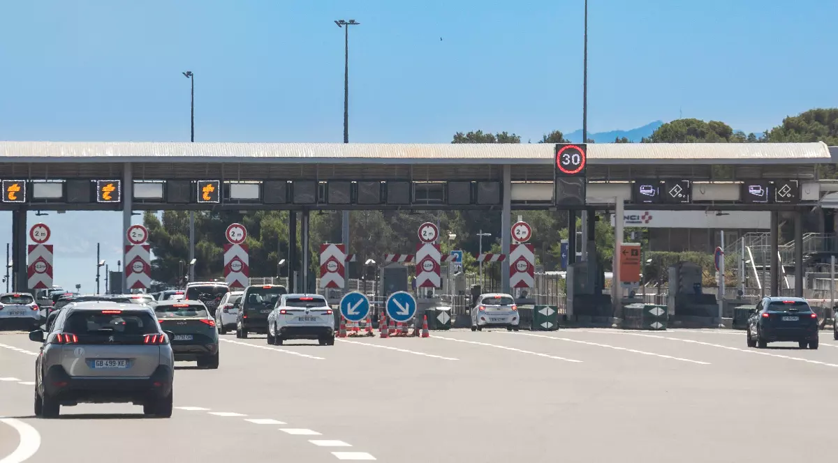 Imaginea prezintă mai m ulte mașini care se apropie sau staționează la barierele de taxare pe o autostradă, cu mai multe benzi ide circulație