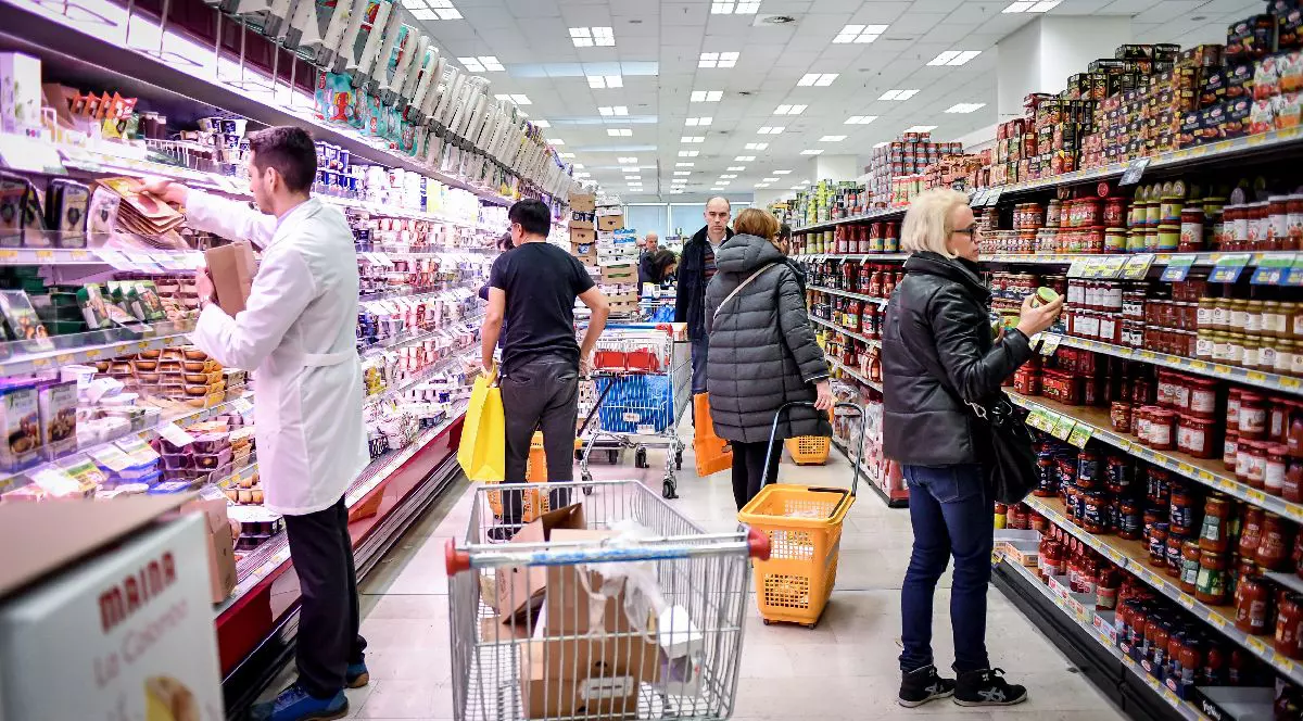 Imaginea prezintă interiorul unui supermarket aglomerat, unde mai mulți clienți analizează produsele de pe rafturi, în timp ce un angajat în halat alb aranjează marfa în secțiunea frigorifică.