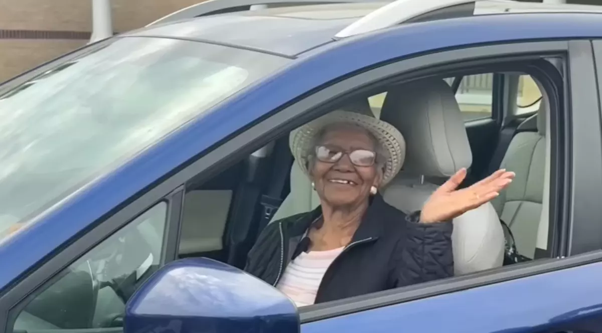 O femeie în vârstă se află la volanul unei mașini albastre și salută zâmbind prin geamul deschis al portierei. Imaginea transmite energie și optimism, sugerând independența și mobilitatea de care continuă să se bucure în ciuda vârstei înaintate.