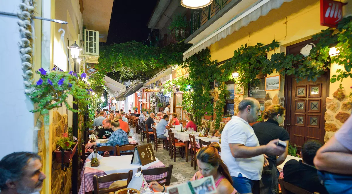 O tavernă grecească animată, plină de turiști, surprinde atmosfera de vacanță din serile de vară din Thassos. Mesele așezate pe aleea îngustă, luminile calde și decorul cu viță-de-vie completează imaginea specifică insulelor grecești.