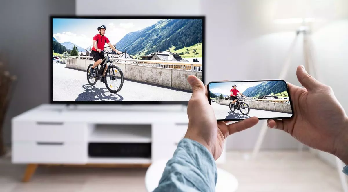 Un bărbat ține în mână un telefon și face o poză de la televizorul deschis, unse vede un biciclist în tricou roșu
