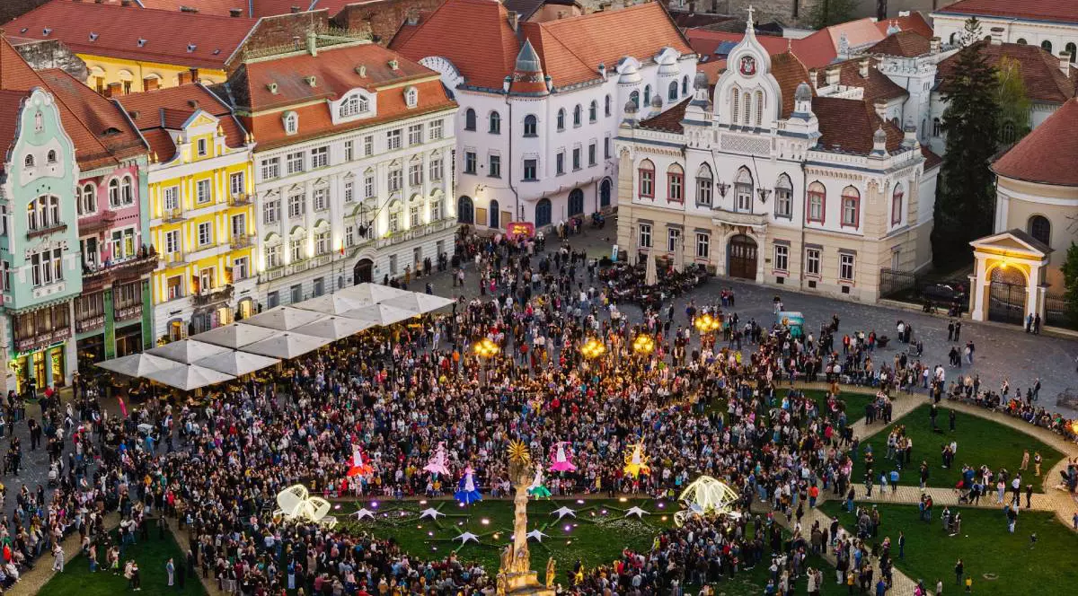 Imagine aeriană din Piața Unirii din Timișoara, unde sute de oameni sunt adunați în fața clădirilor istorice colorate. În prim-plan se vede zona verde a pieței, iar în jur apar fațade baroce, acoperișuri roșii și clădiri emblematice din centrul orașului.