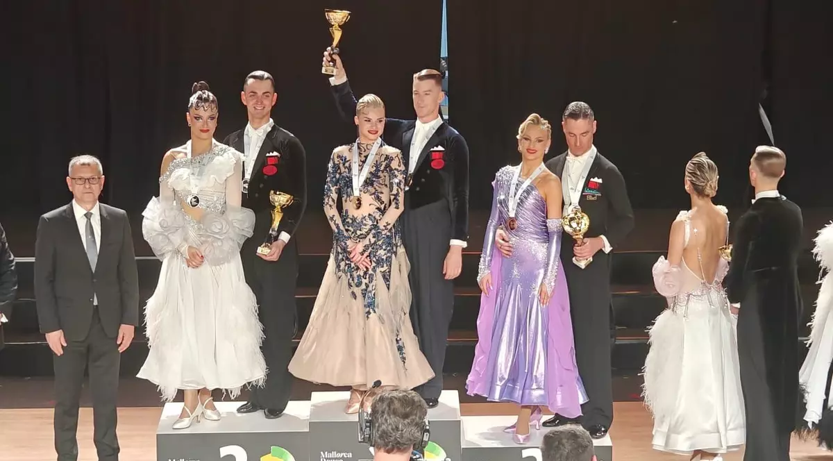 O fotografie de la ceremonia de premiere a unui concurs de dans sportiv, unde trei perechi de finaliști, îmbrăcate elegant în costume de competiție și rochii spectaculoase, stau pe podium alături de un oficial, în timp ce perechea câștigătoare ridică trofeul, totul desfășurându-se pe o scenă cu parchet și un fundal negru, în fața unor cameramani.