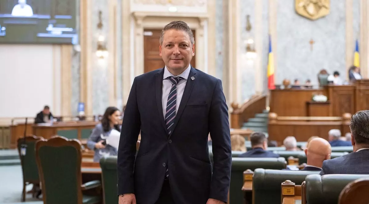 Marius Hudelmicu îmbrăcat într-un costum închis la culoare, cămașă bleu și cravată în dungi.