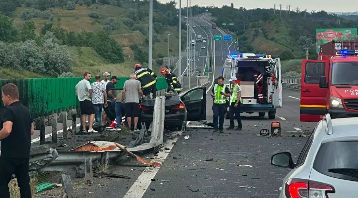 Imaginea surprinde scena unui accident rutier grav produs pe autostrada Soarelui A2, într-o zonă de pod sau viaduct flancată de dealuri cu vegetație. În centrul cadrului, un autoturism puternic avariat este oprit de-a curmezișul benzii, lipit de un parapet metalic separator care este complet deformat și rupt în urma impactului, iar pe asfalt sunt împrăștiate numeroase resturi. În jurul mașinii distruse s-au strâns mai mulți bărbați în haine civile, alături de care intervin direct pe vehicul doi pompieri echipați cu căști și costume de protecție. Pe banda din dreapta este oprită o ambulanță cu ușile din spate larg deschise, în apropierea căreia stau două persoane în uniforme și veste reflectorizante, iar pe jos, lângă salvare, a fost așezat un echipament tehnic portabil cu motor, probabil pentru descarcerare. În extremitatea dreaptă a imaginii se observă parțial o autospecială de pompieri de mari dimensiuni, cu portiera deschisă. În prim-plan, în colțul din dreapta jos, apare spatele unui alt autoturism oprit, pe marginea stângă a sensului de mers se află un panou înalt de protecție, iar șoseaua se continuă în fundal cu o porțiune lungă în rampă, mărginită de stâlpi de iluminat, pe care circulă alte vehicule aflate la distanță, 12 iunie 2026