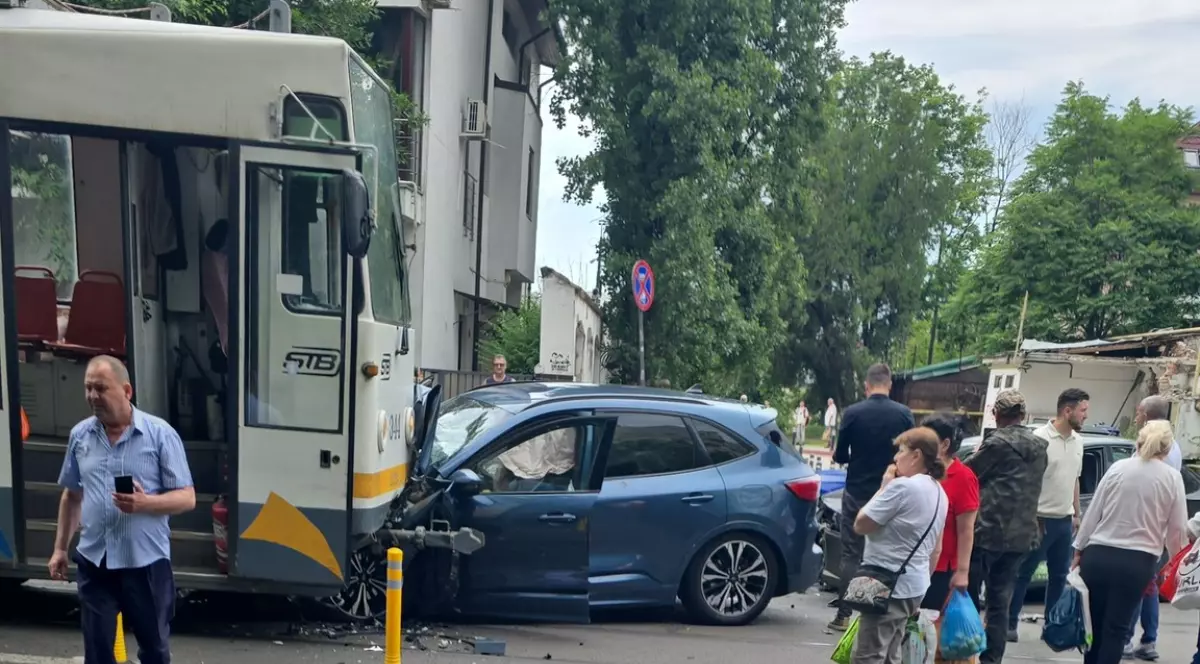 Imaginea surprinde urmările unui accident rutier în care un autoturism albastru a intrat în coliziune cu un vehicul STB, avariile fiind vizibile în partea frontală a mașinii. La fața locului se află mai multe persoane și trecători care privesc scena, în timp ce traficul pare să fie afectat de in