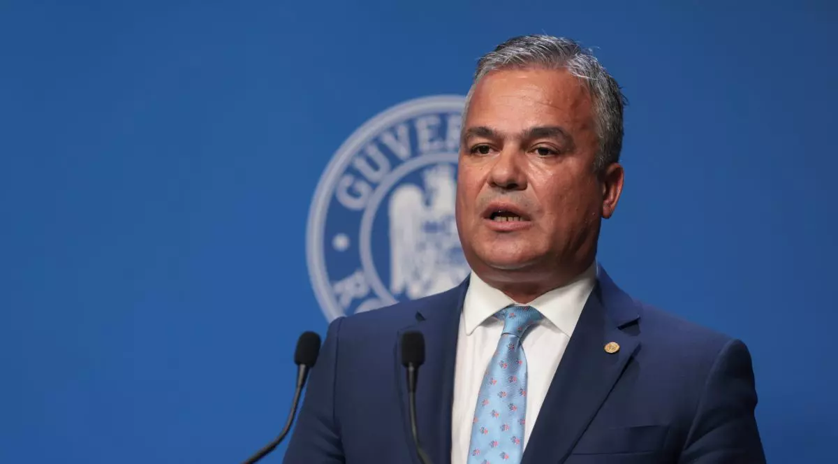 Imaginea prezintă un bărbat îmbrăcat într-un costum închis la culoare și cămașă albă, cu o cravată bleu deschis, surprins într-un cadru oficial. Expresia serioasă și postura sobră sugerează participarea la un eveniment instituțional sau politic.