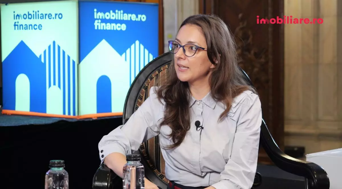 Alexandra Călin de la VivaCity Residence, la interviurile Imobiliare.ro, îmbrăcată cu o cămașă gri, în timp ce stă pe scaun, iar pe fundal se vede logo-ul Imobiliare și Imobiliare.ro Finance