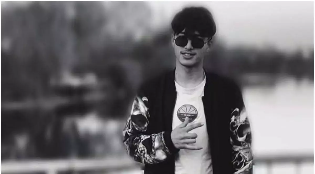 Imaginea alb-negru prezintă portretul unui tânăr purtând ochelari de soare și o jachetă cu model pe mâneci, surprins în timp ce face un gest cu mâna.