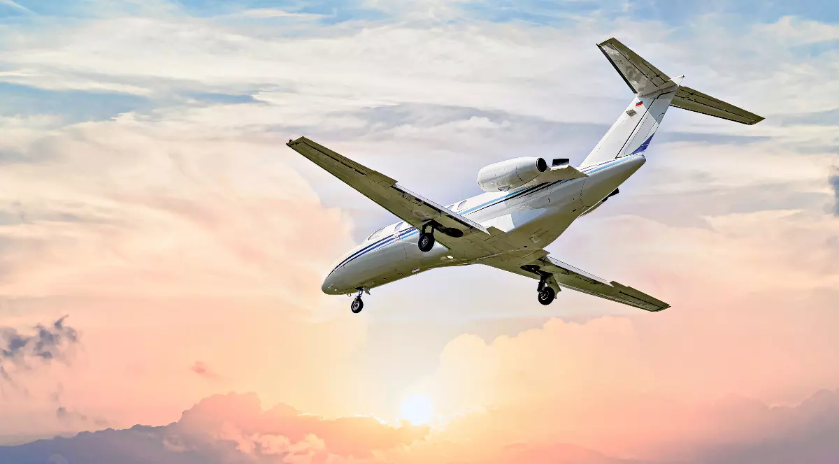 Un avion privat zboară la joasă altitudine pe fundalul unui cer luminat de apus, cu nori colorați în nuanțe de roz și portocaliu. Imaginea ilustrează transportul aerian de lux, adesea asociat cu un nivel ridicat de consum și cu o amprentă de carbon semnificativă.