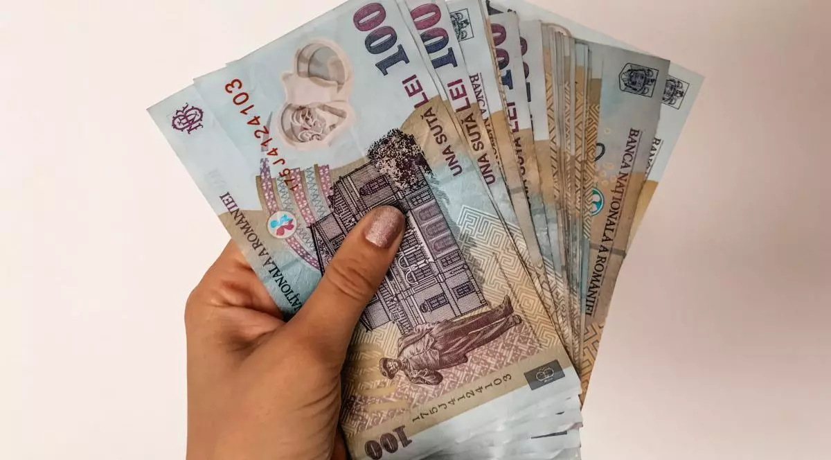 Mâna unei femei, cu unghii făcute cu ojă, care deține mai multe bancnote de 100 de lei, bani românești, pe un fundal bej