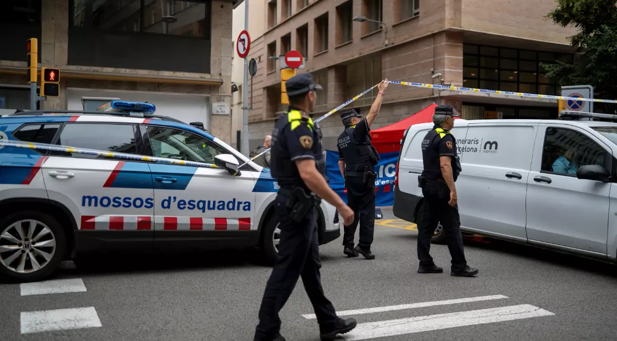 Polițiști care au izolat o zonă stradală cu bandă de securitate în urma unui incident. La fața locului sunt prezenți mai mulți ofițeri în uniformă, o mașină de poliție și o dubă albă de servicii funerare (Serveis Funeraris Barcelona), indicând gestionarea unei scene de crimă.
