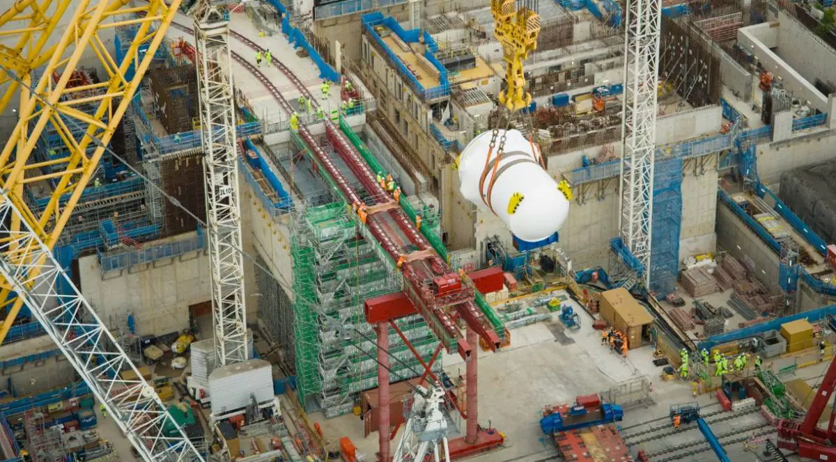 Imagine de pe șantierul centralei nucleare Hinkley Point C din Marea Britanie. Cea mai mare macara din lume ridică un vas alb, uriaș, de reactor nuclear, suspendat de cabluri groase, deasupra unei zone de construcție pline de schele, macarale și muncitori.