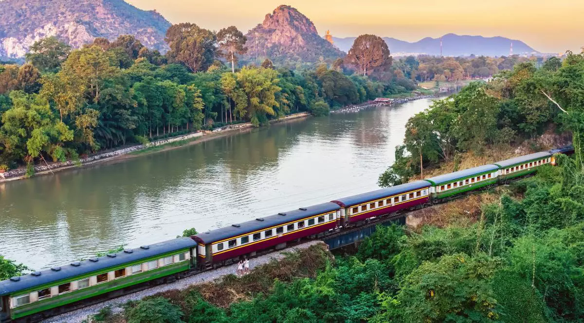 tren de pasageri circulă pe o cale ferată aflată lângă un râu larg, într-o zonă verde și deluroasă, cu multă vegetație, copaci și munți joși în fundal.