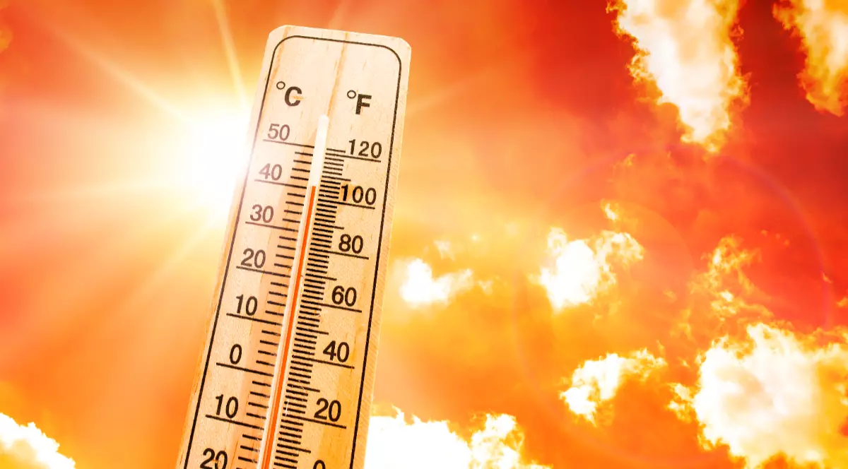 În imagine este prezentat un termometru clasic din lemn care indică o temperatură ridicată de aproape 40 de grade Celsius, fiind plasat pe fundalul unui cer incandescent în nuanțe aprinse de portocaliu și roșu, cu un soare puternic și strălucitor.